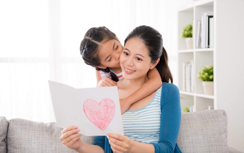 cute little daughter kiss the woman's cheek when the mother received the love card gift and reading happily sitting on the sofa in the living room in mother's day at home.